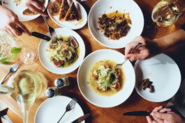 Overhead view of hands and small plates across a shared wooden table with white wine
